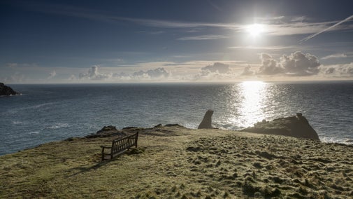 An empty bench near the edge of a cliff with blue sea and skies in the background behind a rocky point